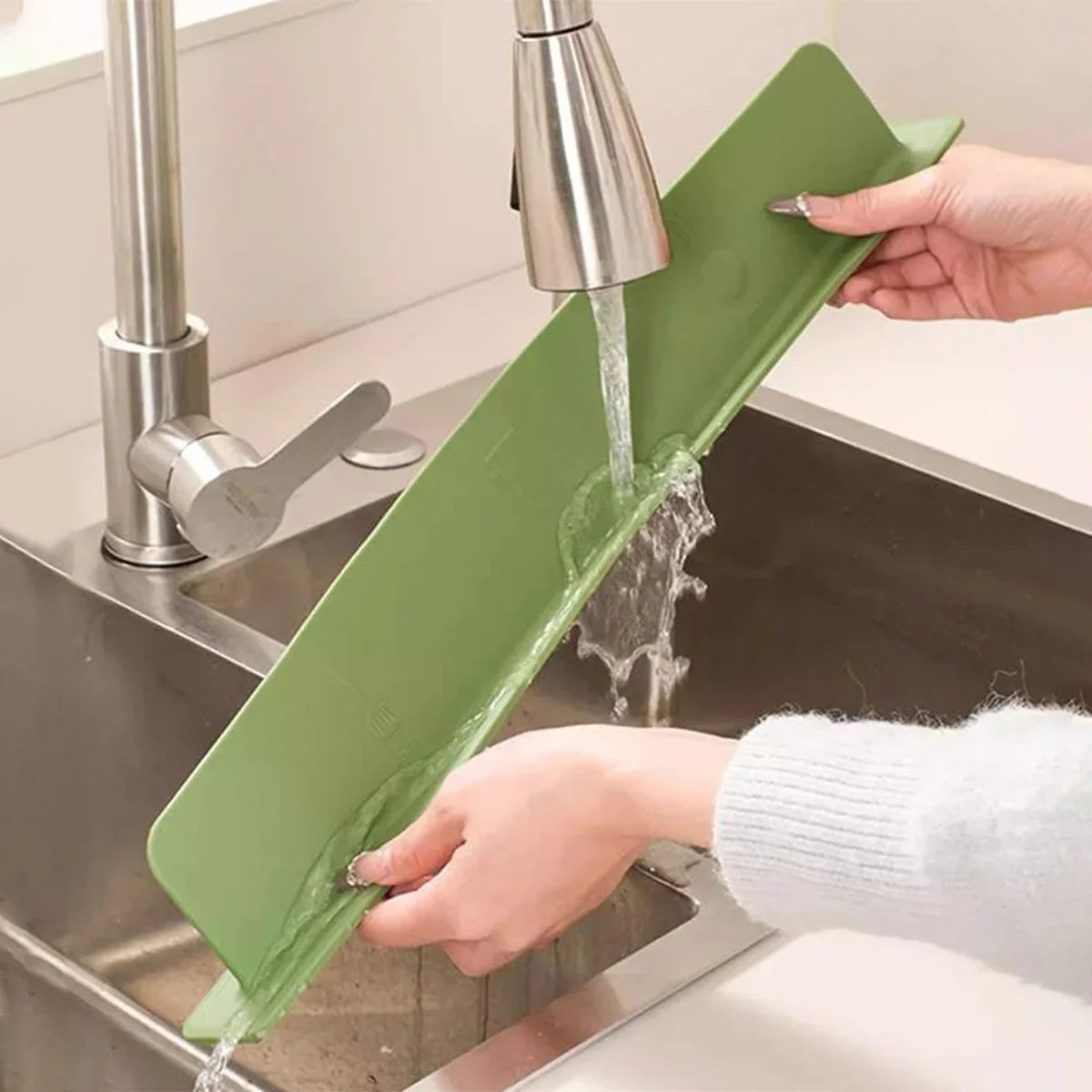 Person washing a green cutting board under running water in a kitchen sink.